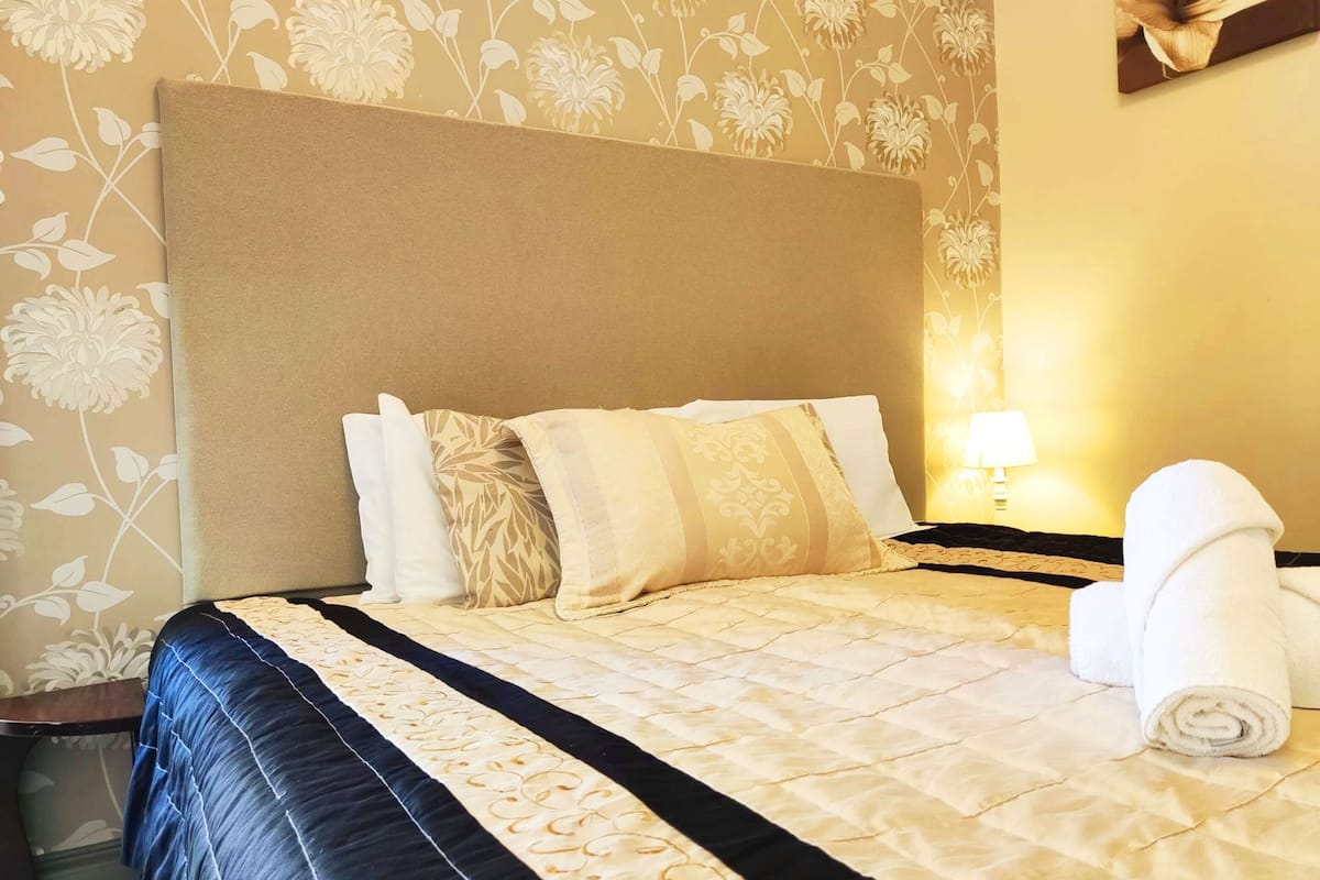 A neatly made bed with a quilted headboard, multiple pillows, and a rolled white towel on the side.
