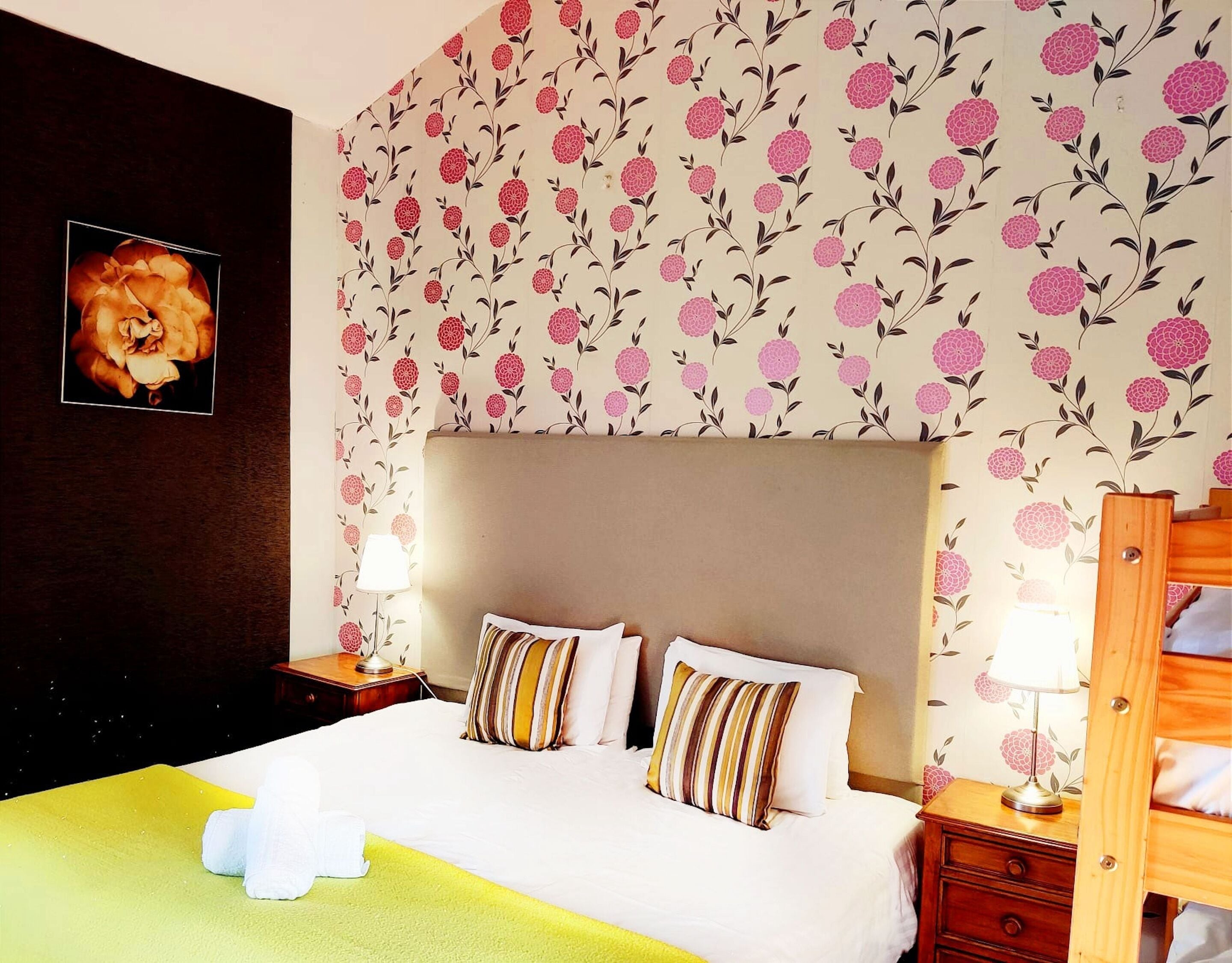 Ein Hotelzimmer mit einem großen Bett, zwei Nachttischen, einem hölzernen Kopfteil, blumigem Wandpapier und einem gerahmten Bild an der Wand.