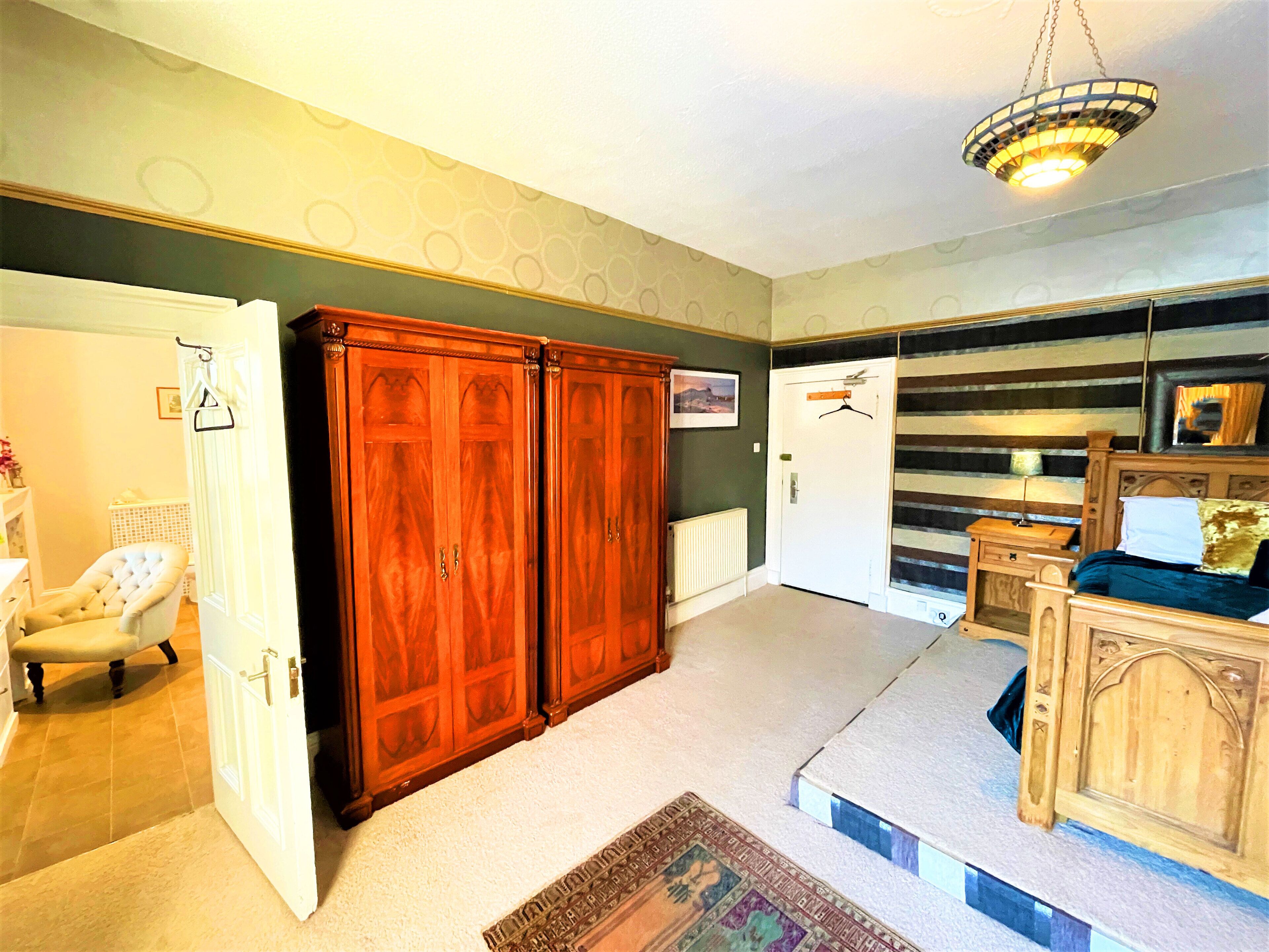 A room with a large wooden wardrobe, a white chair, a bed with a headboard, and a hanging light fixture.