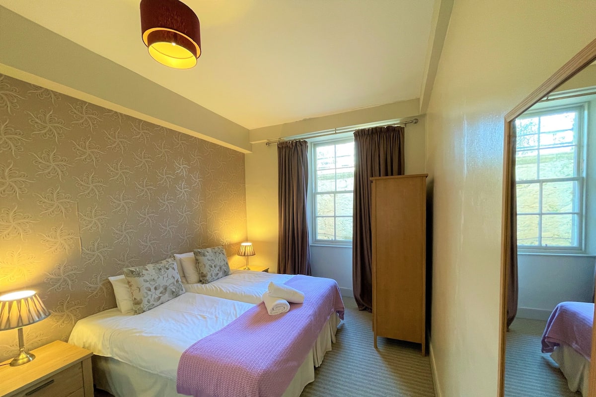 A hotel room with two beds, a wardrobe, a nightstand with a lamp, and a window with curtains.