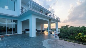 A modern two-story house with a spacious patio area, large windows, and a balcony with potted plants.