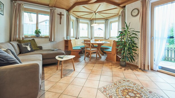 Ein gemütliches Wohnzimmer mit Holzbalkendecke, einer Couch, einem Esstisch mit Stühlen und einem großen Fenster mit Vorhängen.