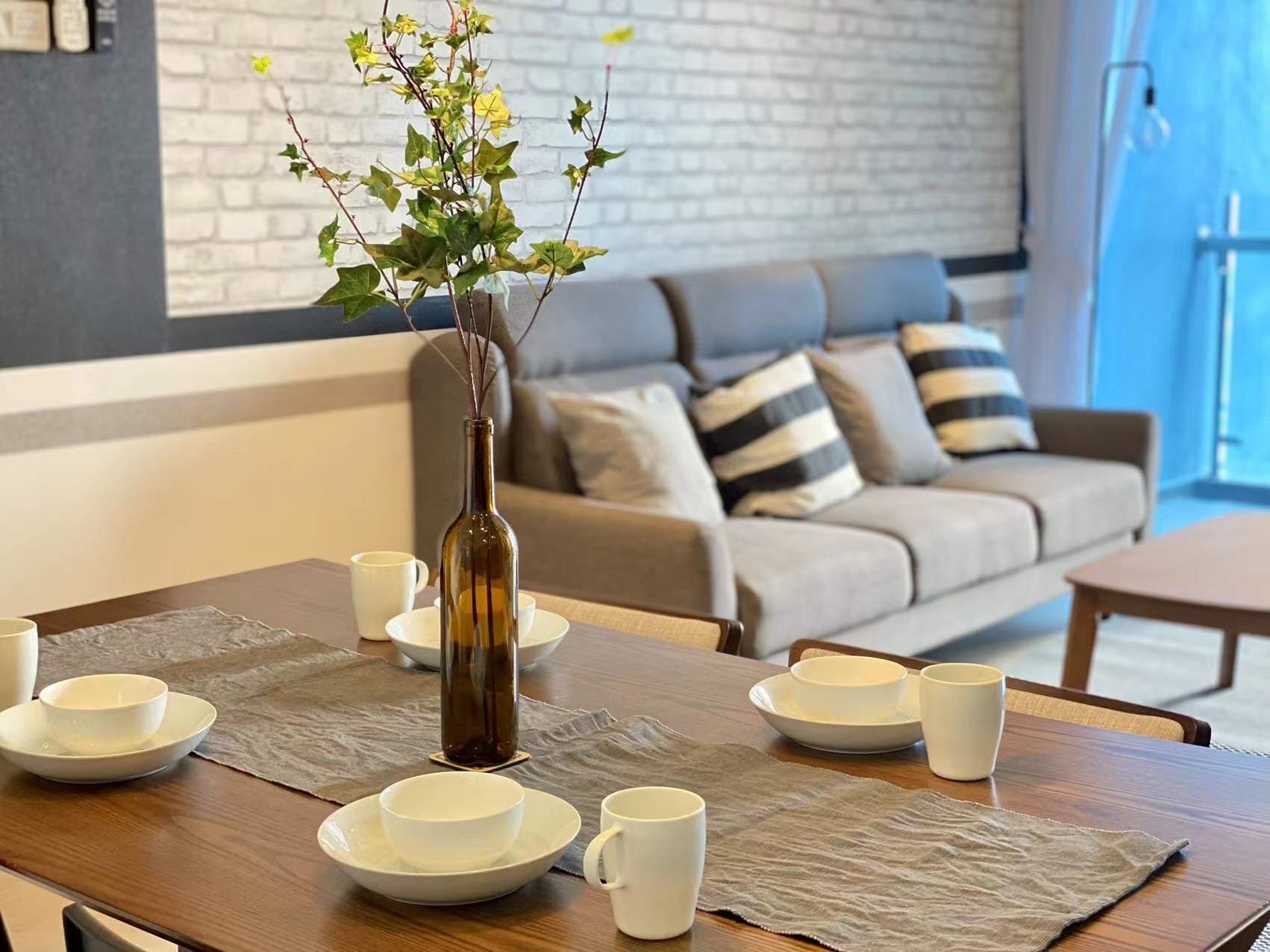 A dining area with a table set for four, a vase with greenery, and a sofa in the background.