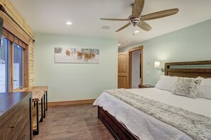 Une chambre à coucher avec un lit, une tête de lit en bois, un ventilateur de plafond, une fenêtre avec des stores et un tableau accroché au mur.