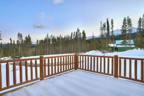 A wooden deck with snow-covered ground, surrounded by tall pine trees and a green building in the background.