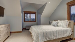 Une chambre avec un plafond en pente, un lit recouvert d’une courtepointe matelassée, une commode en bois et une fenêtre donnant sur des arbres et des montagnes.