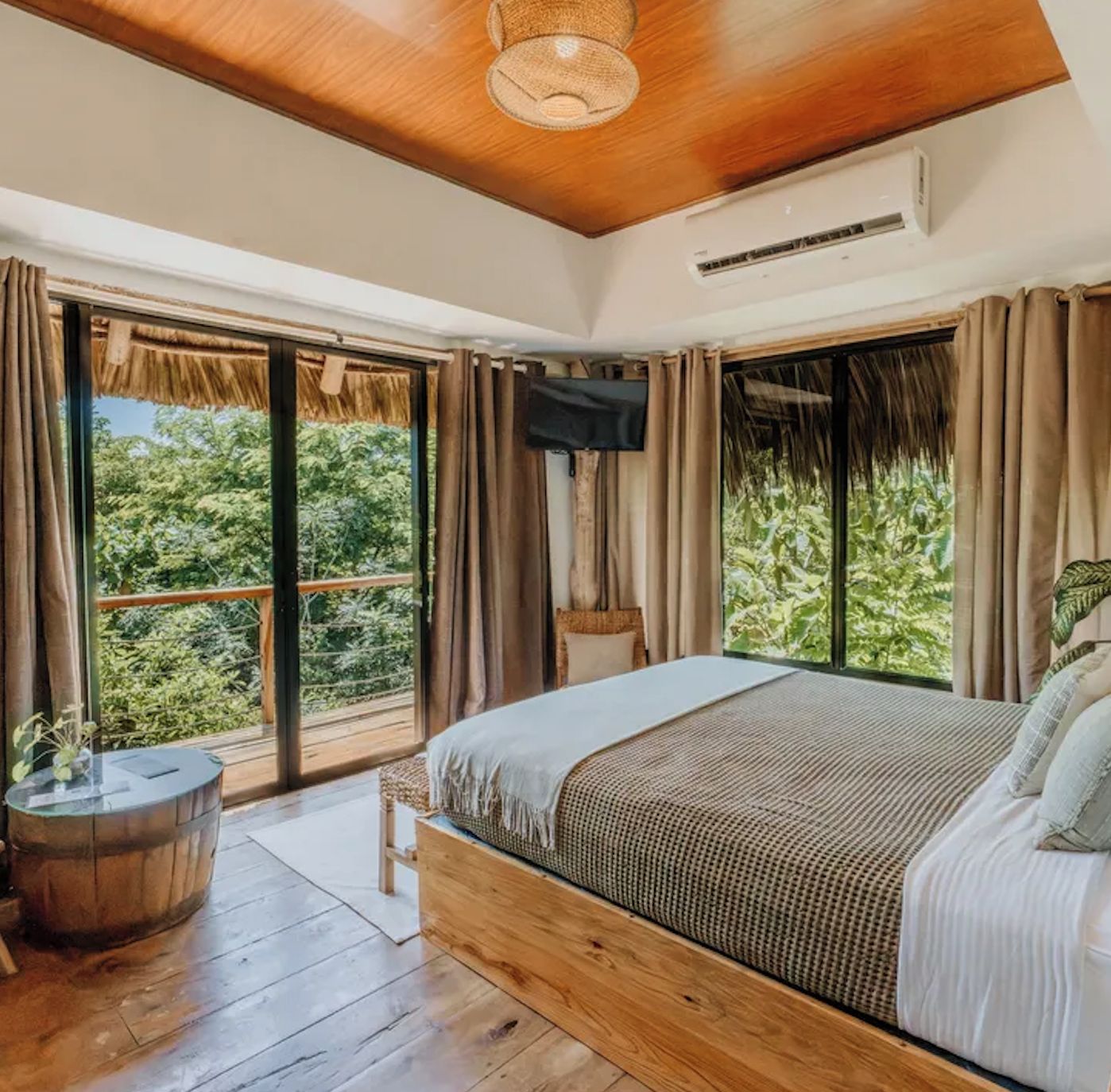 A bedroom with a large bed, wooden flooring, a ceiling fan, and a view of greenery through the windows.