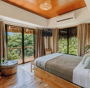 A bedroom with a large bed, wooden flooring, a ceiling fan, and a view of greenery through the windows. - NAIA Miravalle (Sonsonate)