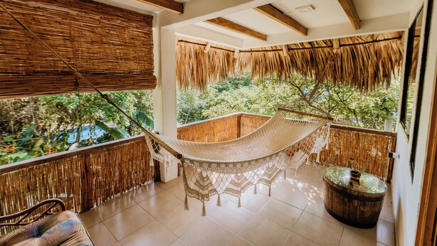 Un hamac sur un balcon donnant sur un paysage verdoyant, une chaise en osier et un tonneau en bois.