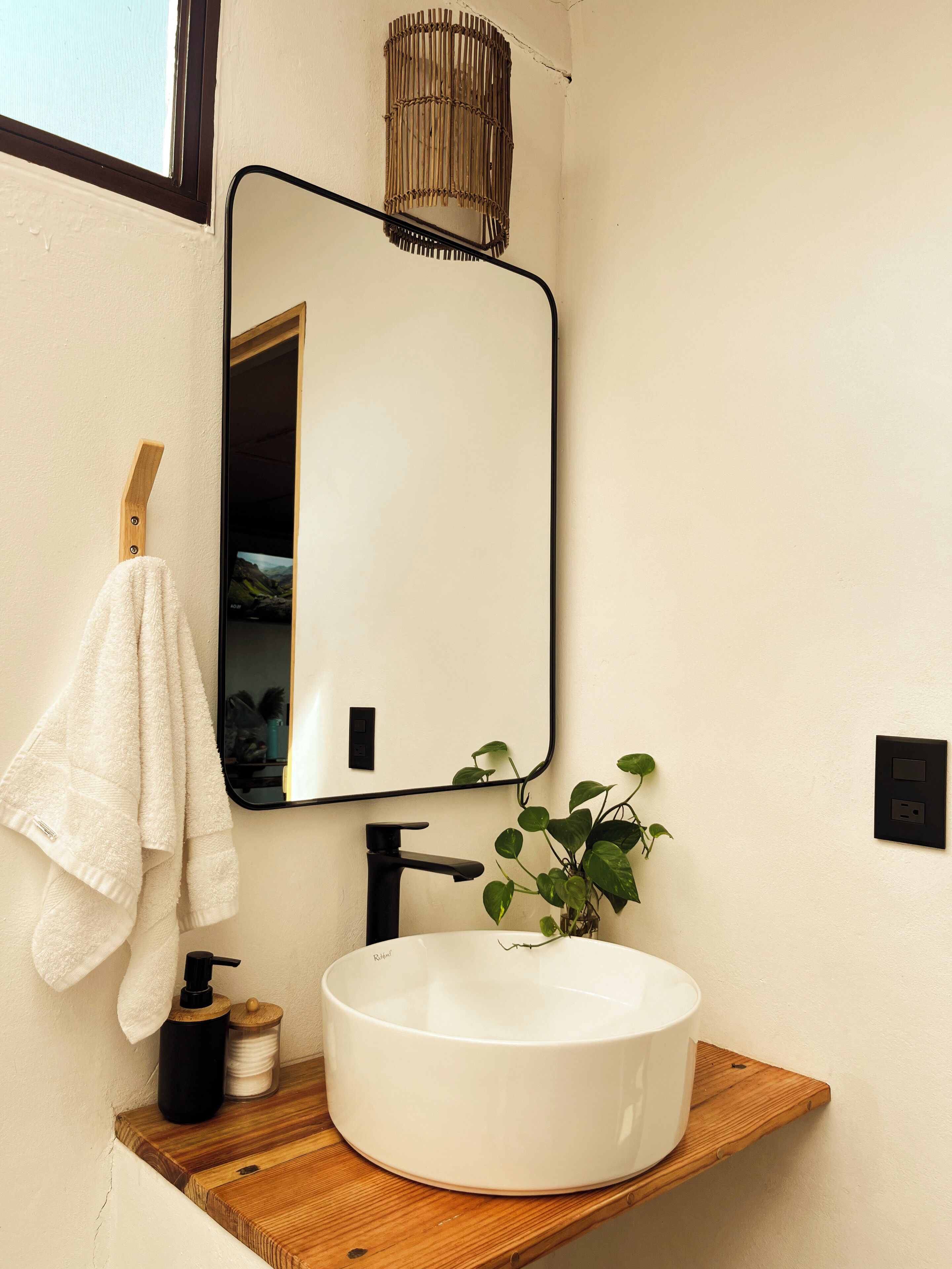 Un baño con un lavamanos blanco, grifo negro y un espejo encima. Hay una toalla colgada en un toallero de madera, una planta en maceta en el borde del lavamanos y una repisa de madera con una botella y un recipiente.