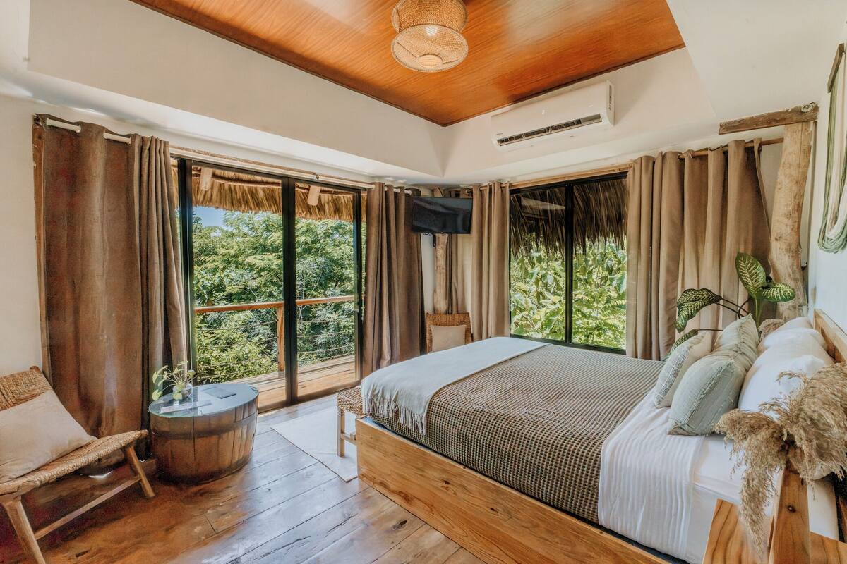 A bedroom with a large bed, wooden flooring, a TV, and a balcony with curtains.