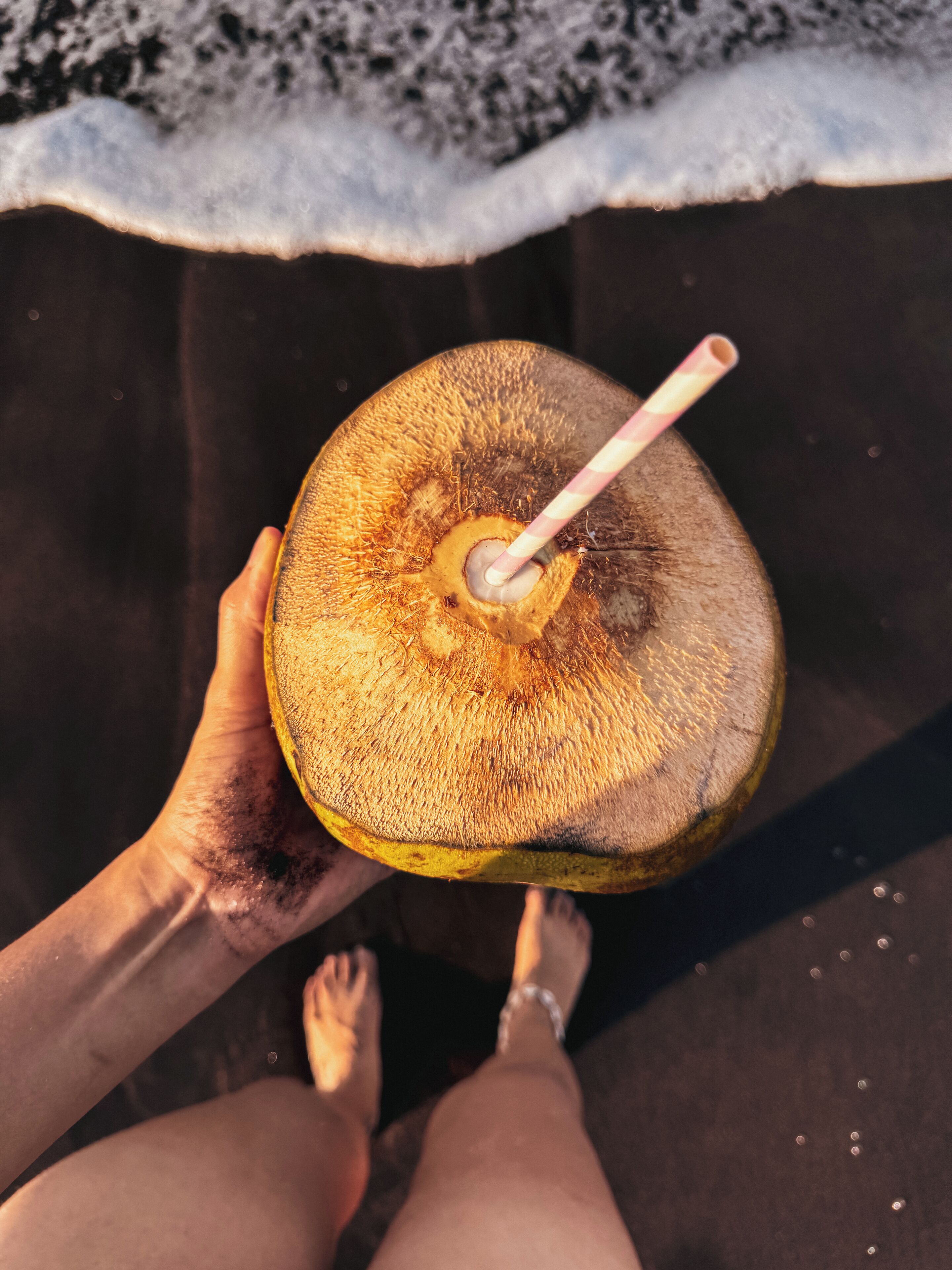 A person holding a coconut with a straw, standing on a sandy beach with waves in the background.