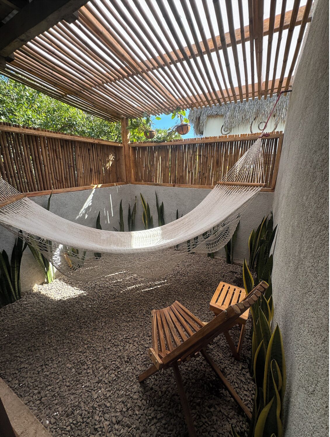 A hammock under a wooden pergola, surrounded by pebbles and plants, with a wooden bench and a thatched roof structure in the background.