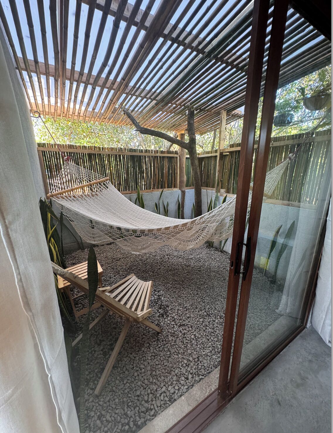 A hammock under a bamboo pergola, a wooden chair, and a glass door leading outside.
