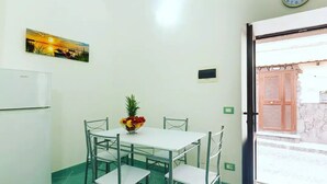 A small, clean room with a white table, four chairs, a bowl of fruit, a clock, and a window with a view of a building. - 1 Bedroom Apartment in Ischia (Ischia)