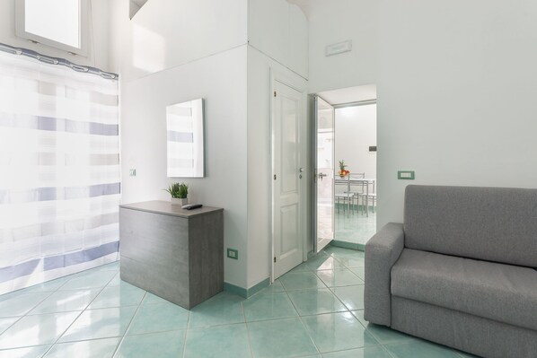 A modern interior with a reception desk, a grey sofa, and a view into a dining area. - 1-Bedroom Apartment in Ischia (Ischia)