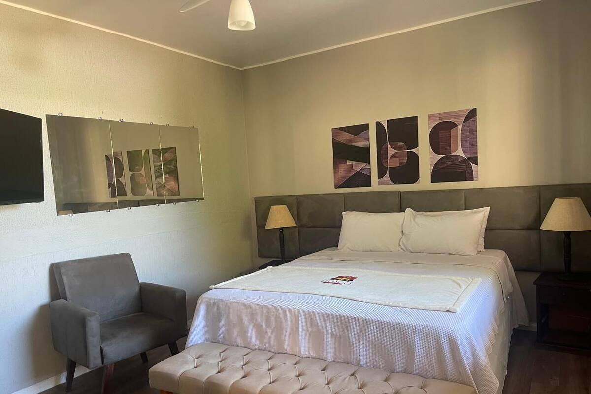 A bedroom with a bed, two bedside lamps, a chair, a bench, and abstract wall art.