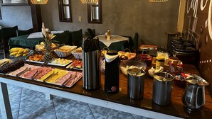 Una mesa de buffet con diversos alimentos y utensilios de cocina, incluyendo una cafetera y ollas, en una sala con mobiliario de madera y luces colgantes.