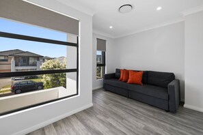 A modern living room with a grey sofa, a window overlooking a residential area, and a ceiling light fixture. - Your Home Away from Home: 4BR Escape (Marsden Park)