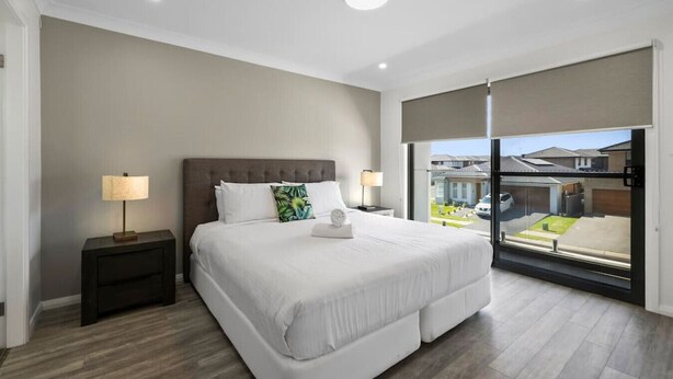 Une chambre d’hôtel moderne avec un grand lit, deux tables de chevet et une vue sur un quartier résidentiel, accessible par une porte coulissante.