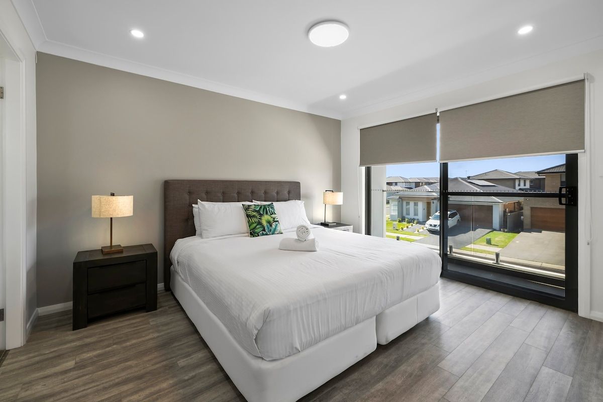 Een moderne slaapkamer met een groot bed, twee nachtkastjes en uitzicht op een woongebied door een schuifdeur.