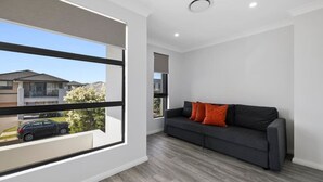 Un salon moderne avec un canapé gris, une grande fenêtre et une vue sur une rue résidentielle.