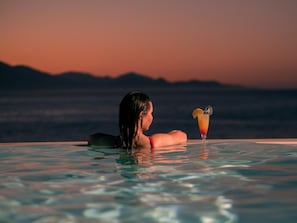 Una persona relajándose en una piscina con una bebida, con vistas a un paisaje montañoso.