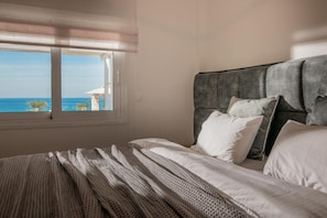 Un dormitorio con un gran ventanal, una cama con ropa de cama blanca y un cabecero tapizado en gris.
