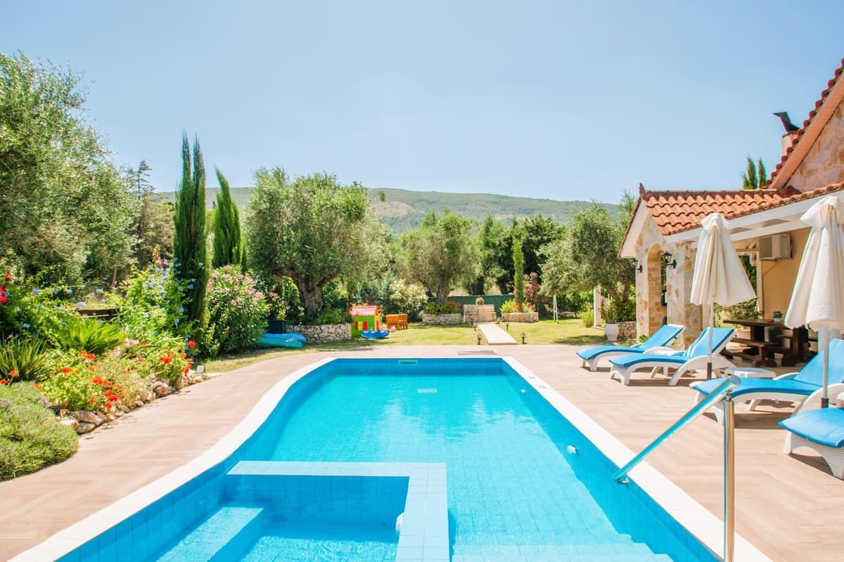 A swimming pool with lounge chairs and umbrellas, surrounded by a garden with various plants and a small play area.