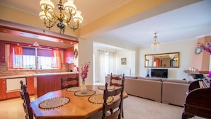 A spacious living area with a dining table, chairs, and a sofa. There is a kitchen area with a window and a chandelier. - Zante Sun II - Getaway Villa! (Zakynthos)