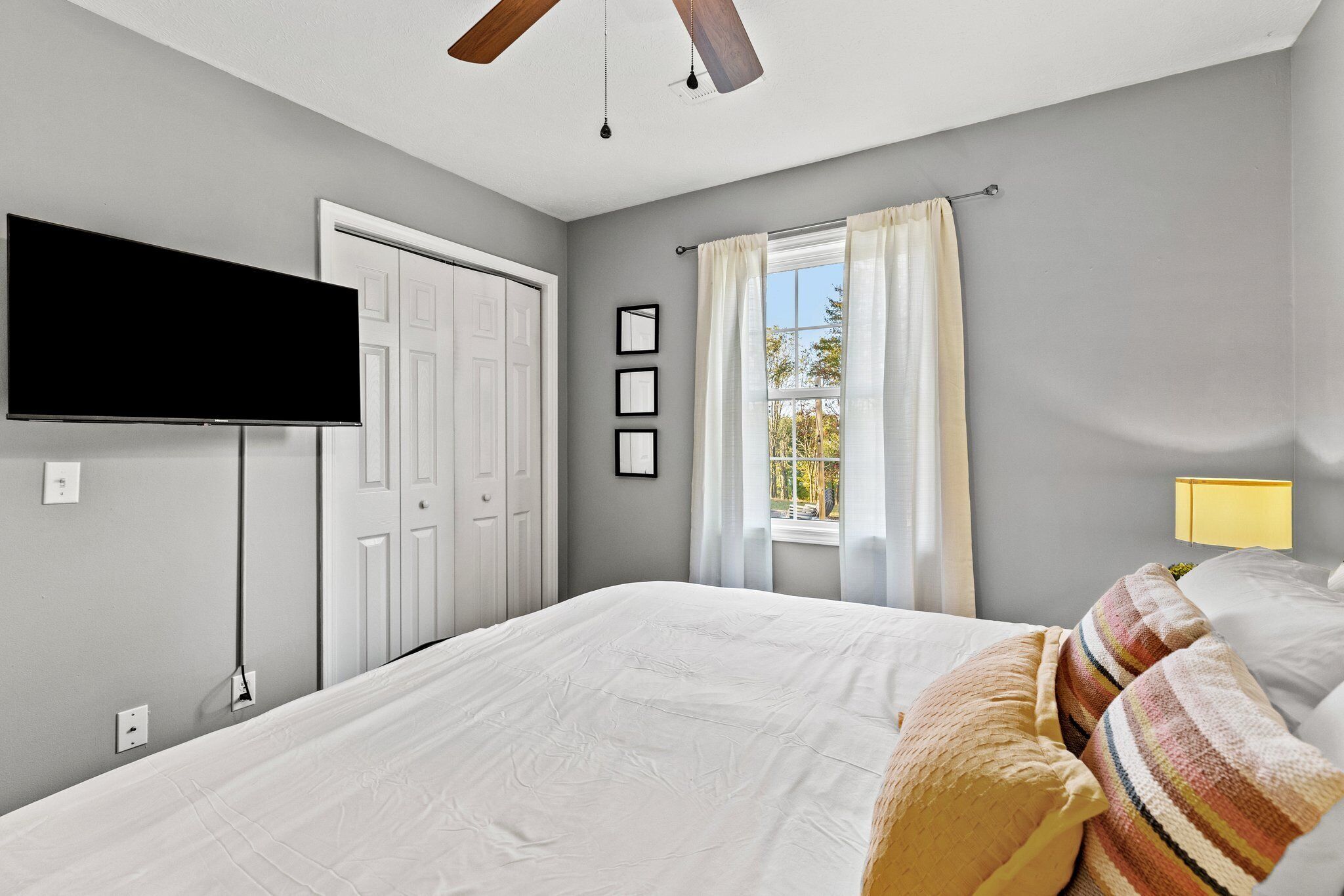A bedroom with a bed, a television mounted on the wall, a window with curtains, and a ceiling fan.