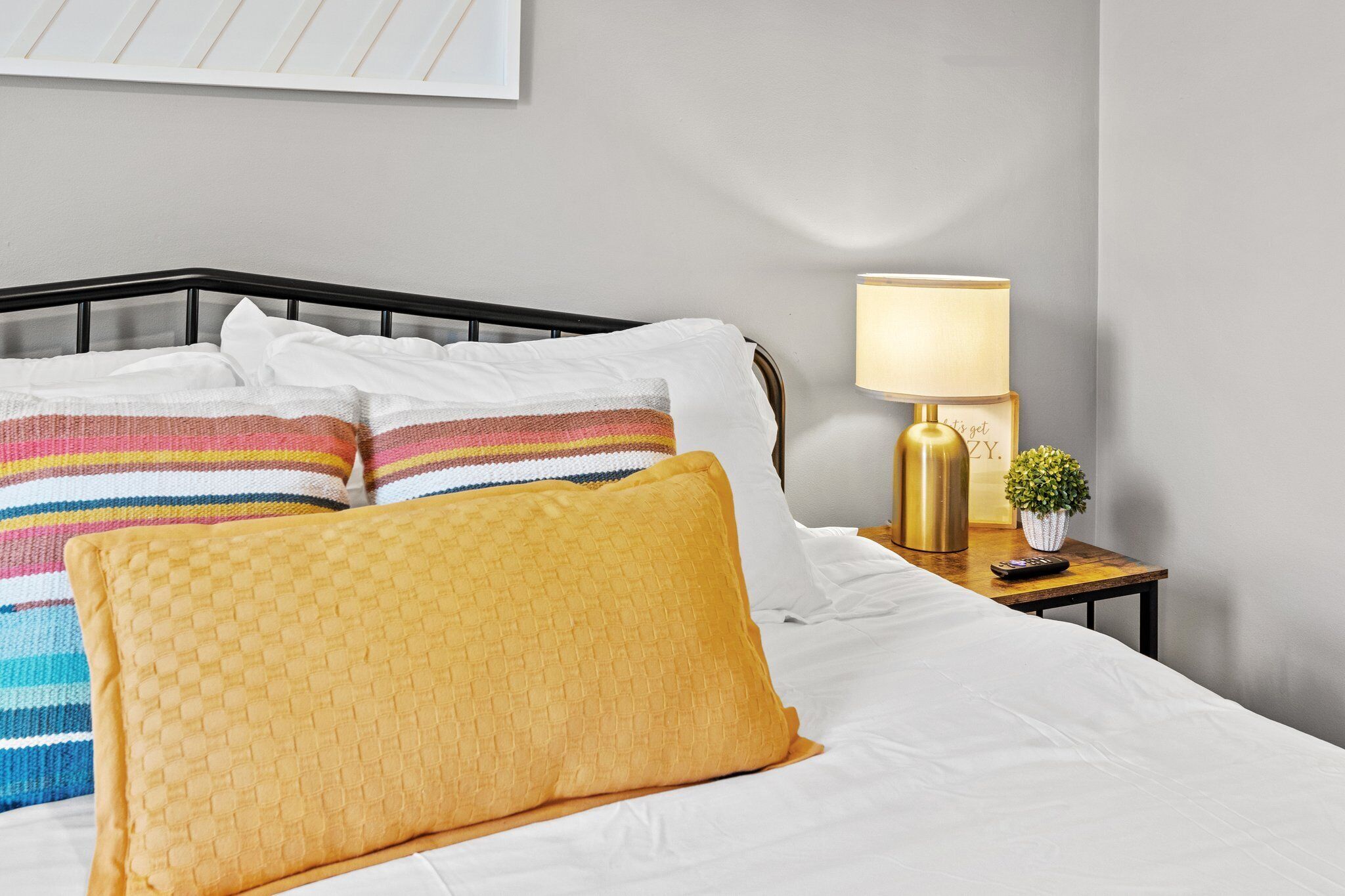 A neatly made bed with white linens and colorful striped pillows, a bedside table with a lamp, and a small potted plant.