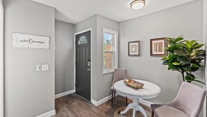 Een modern interieur met een grijze muur, een welkomstbord, een donkere deur, een raam, een ronde tafel met een mand, twee stoelen en een kamerplant.