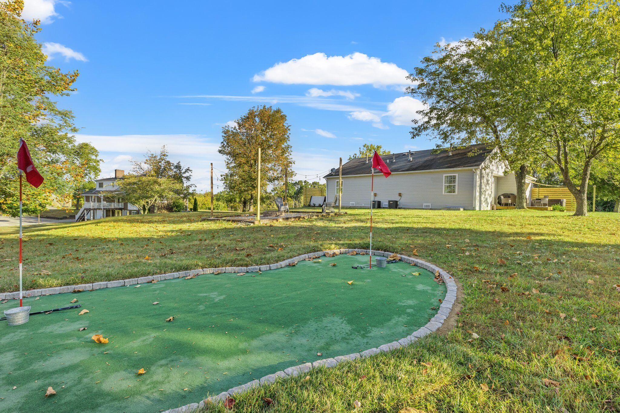 A miniature golf course with a green putting surface, surrounded by a stone border, trees, and a house in the background.