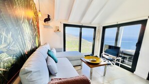 A modern living room with a large window offering a scenic view, a sofa with cushions, a coffee table, and a mounted artwork. - Mar a Vista by Your Madeira Rentals (Ponta do Sol)