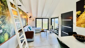 A modern living room with a white sofa, a coffee table, and a dining table. There is a ladder against the wall, a ceiling fan, and large windows offering a view of the outdoors. - Mar a Vista by Your Madeira Rentals (Ponta do Sol)