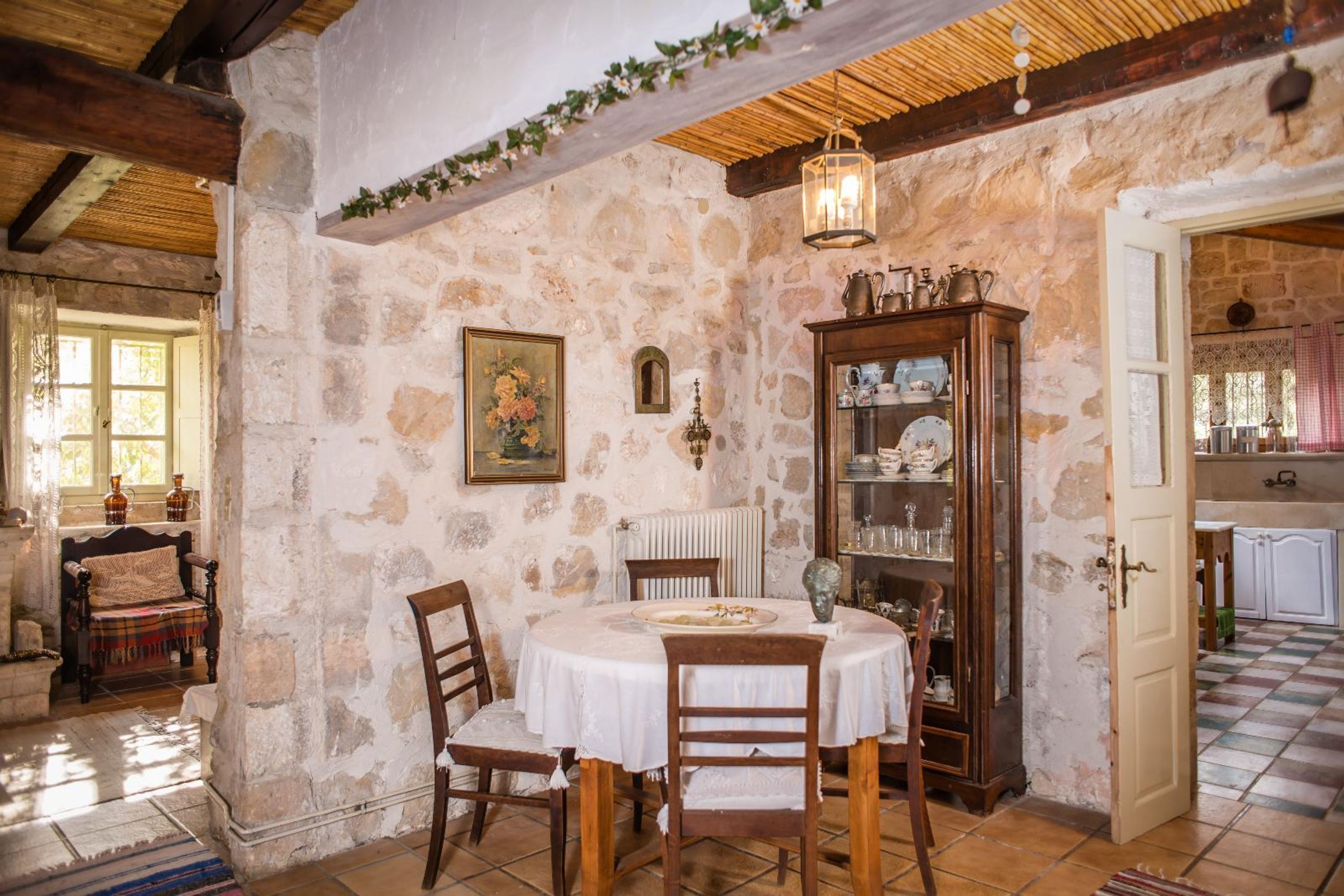 Une pièce rustique aux murs de pierre, avec une table ronde, des chaises en bois, un buffet contenant de la verrerie et une fenêtre ornée de rideaux.