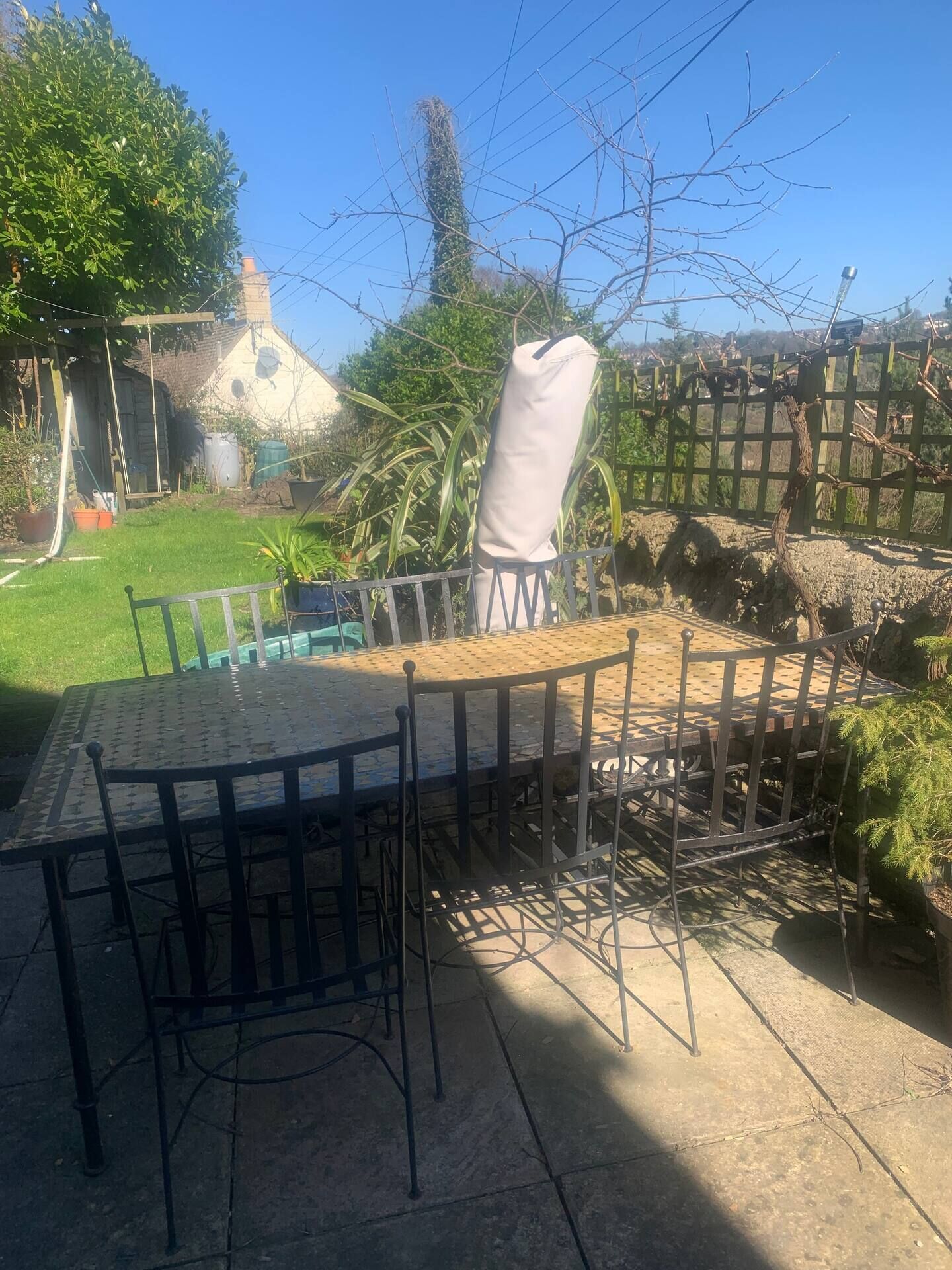 A round metal table with chairs, a concrete patio, and a green fence in a garden setting.