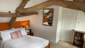 A bedroom with a wooden ceiling beam, a white bed with white and striped pillows, a bedside table with a lamp, a mirror, and a bookshelf. - Traditional Cotswold cottage with stunning views and walking (Stroud)