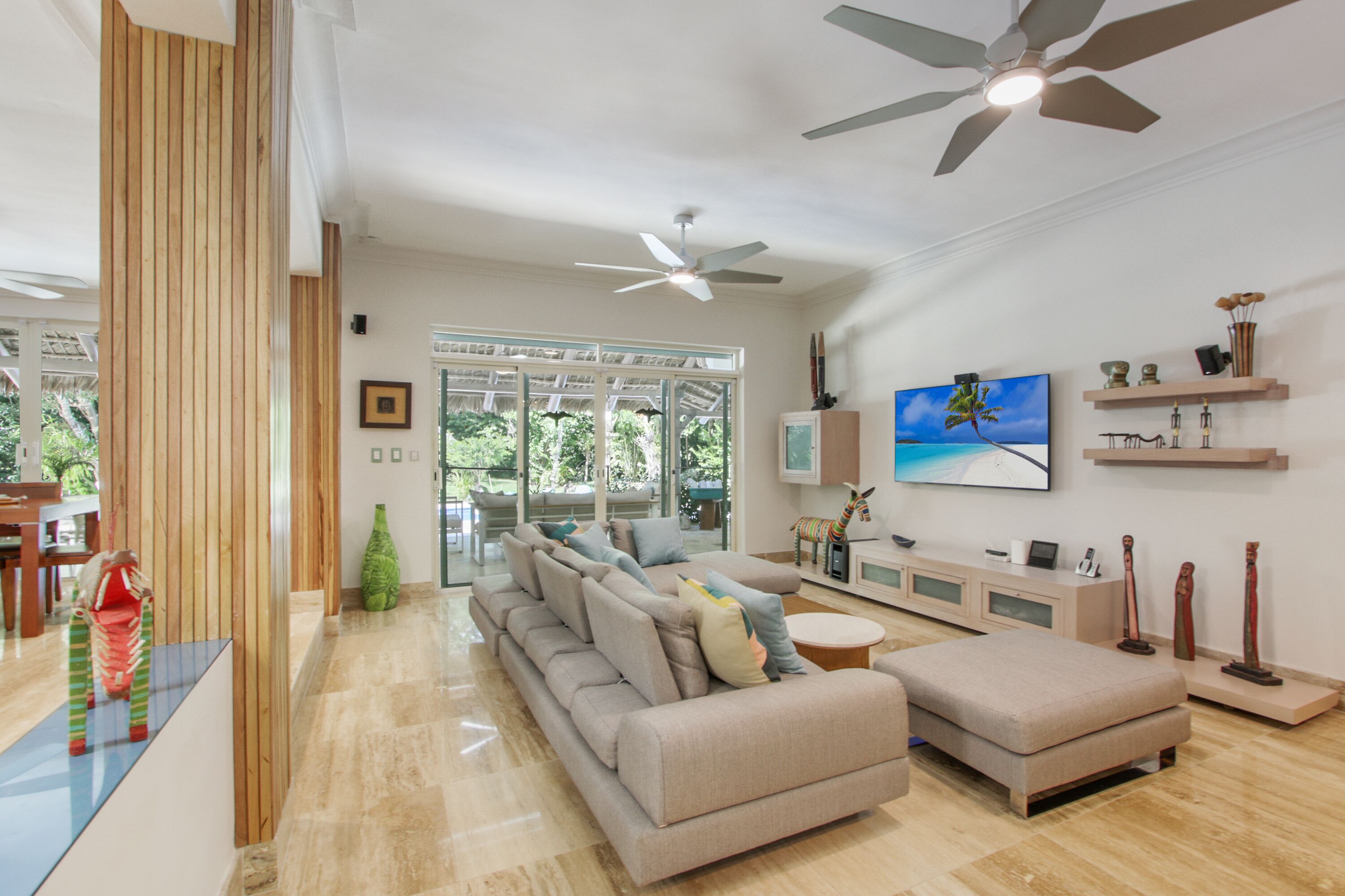 A modern living room with a beige sofa, a coffee table, and a TV mounted on the wall. There are ceiling fans, a wooden accent wall, and a large window providing natural light.