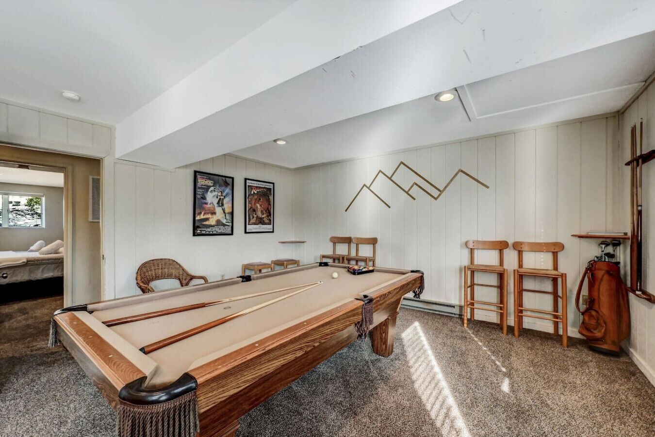 Una mesa de billar en una habitación con paredes blancas y un techo alto. Hay dos sillas de madera y una mesita junto a la mesa de billar.