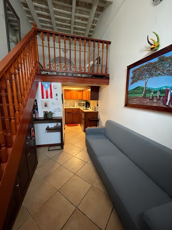 Un vaste espace de vie agrémenté d’un escalier en bois, une cuisine avec des meubles en bois, un canapé gris et un tableau représentant un paysage.