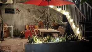 Una escalera con luces cálidas, un paraguas rojo, una mesa, sillas y plantas en macetas.