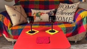 Una mesa roja con dos copas de vino y un mantel rosa, frente a un sofá colorido con un cojín de mapa mundial y un cojín que dice 'HOGAR Dulce HOGAR'.