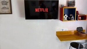 Una habitación con un televisor de montaje, que muestra el logotipo de Netflix, una pequeña mesa de madera con una cesta y un estante con objetos decorativos.