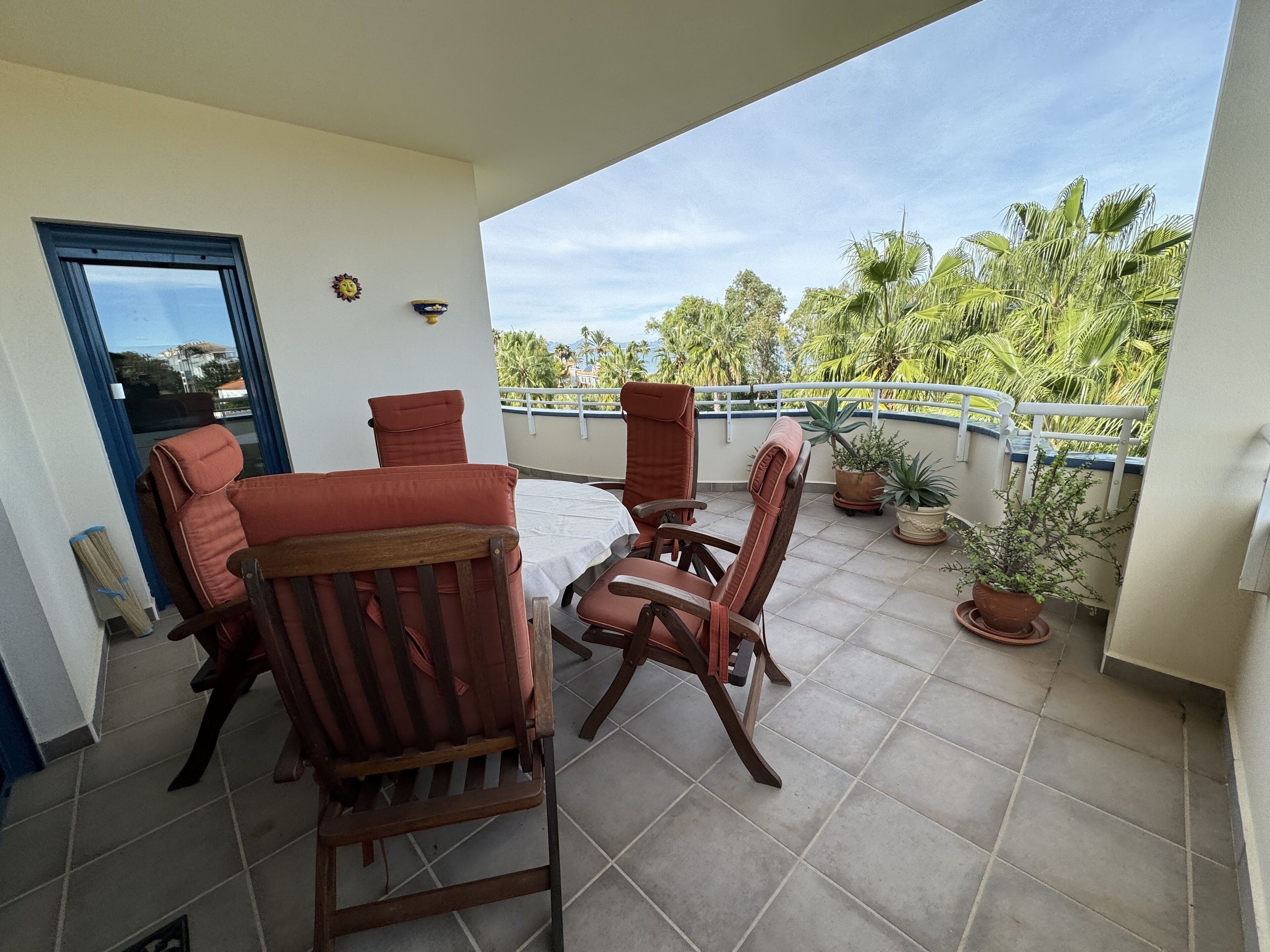 A balcony with two wooden chairs and a table, potted plants, and a view of palm trees and buildings.