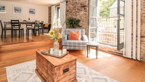 Un salon spacieux doté d’un plancher en bois, d’un canapé à carreaux agrémenté d’un coussin orange, d’une table basse en bois sur laquelle est posé un vase de fleurs, et d’un coin repas avec une table dressée pour quatre personnes.