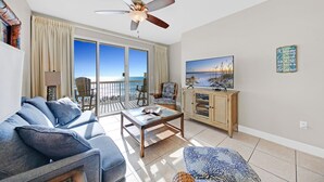 A spacious living room with a large sofa, a coffee table, and a television. There is a balcony with a view of the beach.