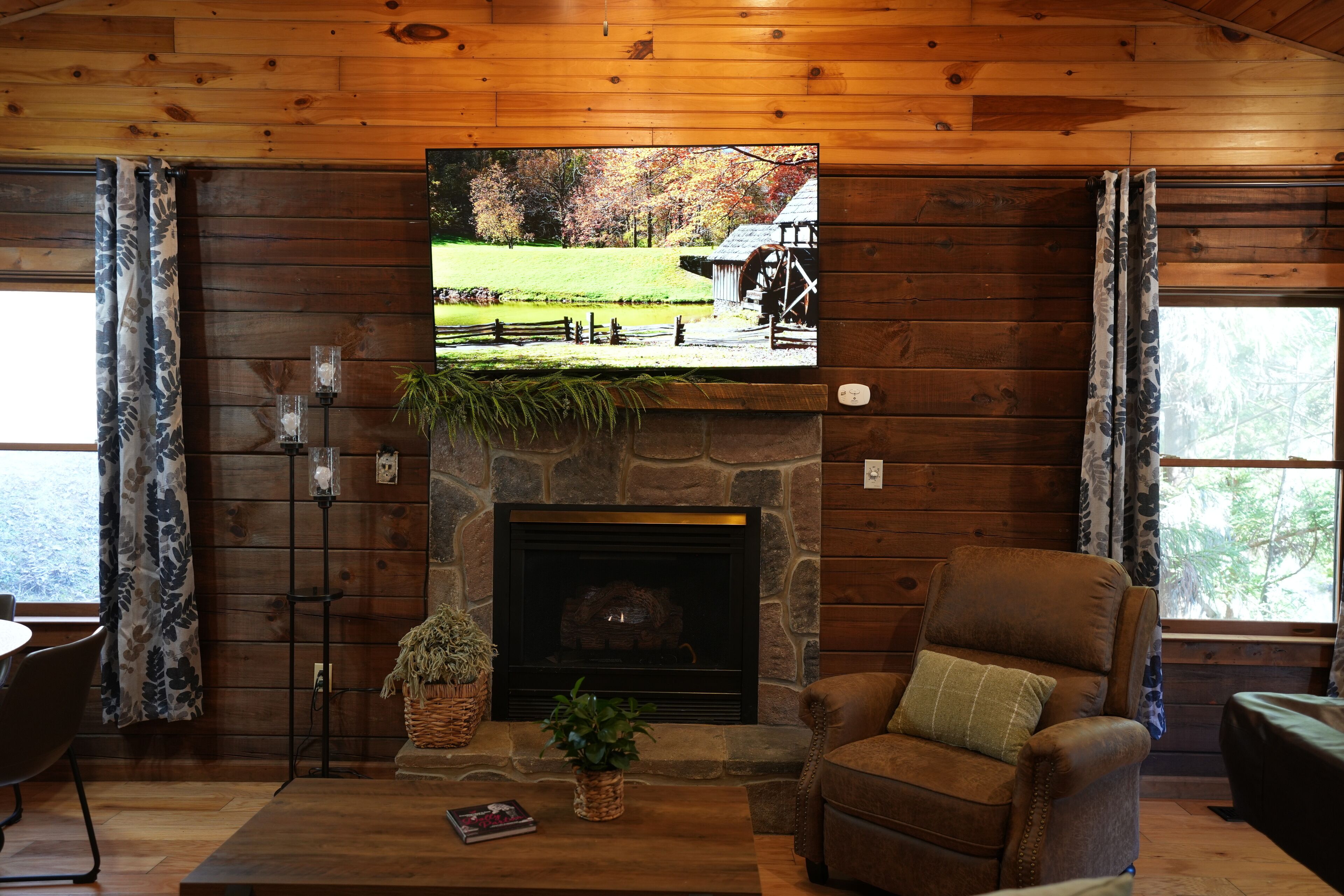 A cozy cabin interior with a stone fireplace, a comfortable leather armchair, and a television mounted above the fireplace displaying a scenic view.