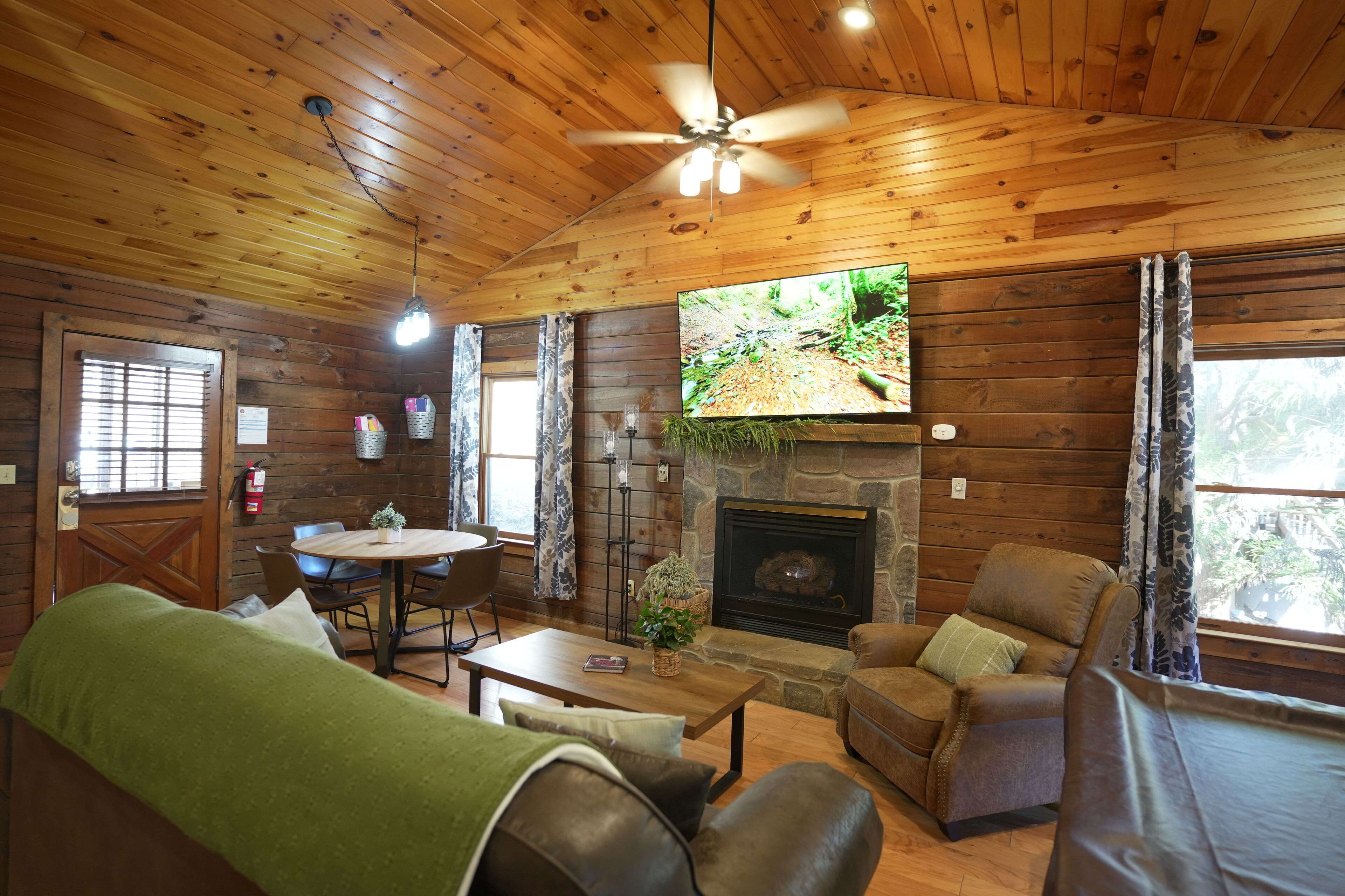 A cozy cabin interior with a stone fireplace, wooden walls, a ceiling fan, and a television mounted above it.